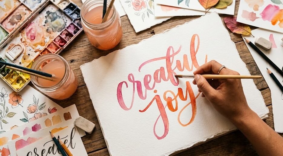Melhor Papel para Lettering Vista superior de um artista pintando um lettering colorido e vibrante em um papel texturizado grosso, cercado por tintas aquarela.