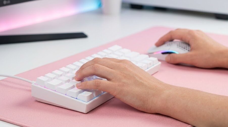 Mãos de uma única jogadora utilizando um teclado mecânico branco e um mouse ultraleve sobre um mousepad rosa estendido.