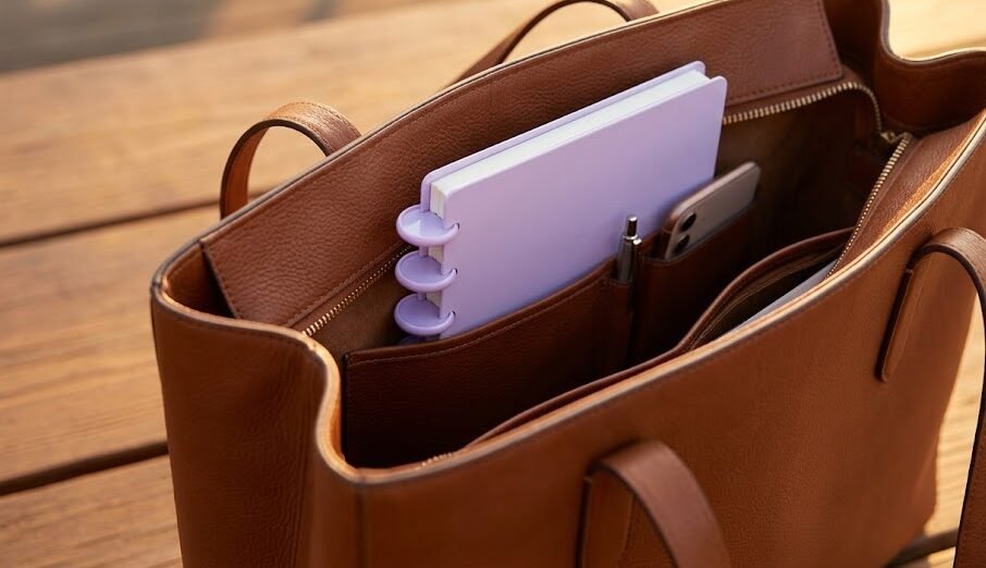 Caderno inteligente tamanho A5 pequeno de cor pastel aparecendo de dentro de uma bolsa de couro feminina moderna.